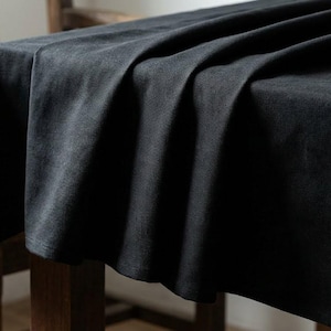May include: A dark charcoal linen tablecloth draped over a wooden table. The fabric has a textured appearance with soft folds and shadows. The table is set near a wooden chair, suggesting a dining or kitchen setting.