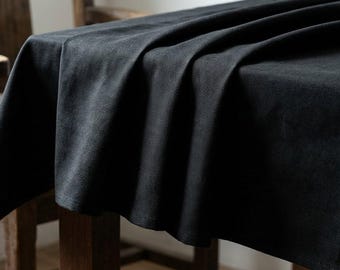 May include: A dark charcoal-colored linen tablecloth draped over a wooden table. The fabric has a textured appearance with soft folds and shadows. The table is set near a wooden chair, suggesting a dining or kitchen setting.