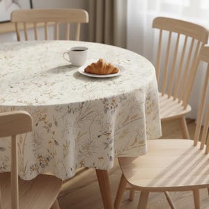 Puede incluir: Un mantel redondo con un estampado floral en beige, verde y amarillo cubre una mesa de madera. Una taza blanca y un plato con un cruas&aacute;n est&aacute;n sobre la mesa. Cuatro sillas de madera rodean la mesa.