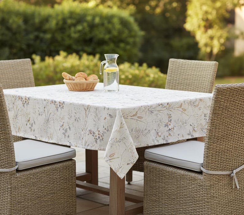 Puede incluir: Un juego de mesa de comedor al aire libre con un mantel floral. Sillas de mimbre con cojines grises rodean la mesa. Una cesta de panecillos y una jarra de agua con rodajas de lim&oacute;n est&aacute;n sobre la mesa.