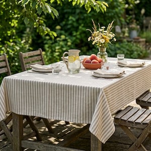 May include: A rectangular wooden outdoor table covered with a beige and white striped tablecloth. The table is set with four white plates, folded napkins, clear glasses, a glass pitcher of lemonade, and a bowl of peaches next to a flower vase.