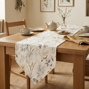 May include: A rectangular table runner featuring a cream-colored fabric with a botanical print of grey and brown foliage, delicate branches, and small birds. The runner is draped over a wooden dining table set with a teapot, cups, and plates.