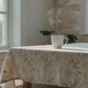 Puede incluir: Una taza blanca humeante sobre una mesa cubierta con un mantel floral. El mantel presenta un estampado de p&aacute;jaros, flores y follaje en tonos beige, gris y amarillo. Luz natural.