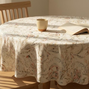 May include: A round tablecloth with a floral pattern in shades of beige, green, and red covers a wooden table. A ceramic mug and an open book rest on the tablecloth. Wooden chairs surround the table, creating a cozy dining setting.