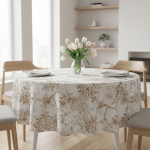 May include: A round tablecloth with a floral pattern in shades of brown and beige covers a table set for dining. Two plates are on the table, and a vase of tulips sits in the centre. Wooden chairs surround the table.