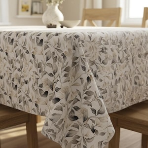 Puede incluir: Un mantel rectangular con un patrón repetido de hojas grises y beige sobre un fondo blanco. El mantel está colocado sobre una mesa de madera. El tema general es la naturaleza y la decoración del hogar.