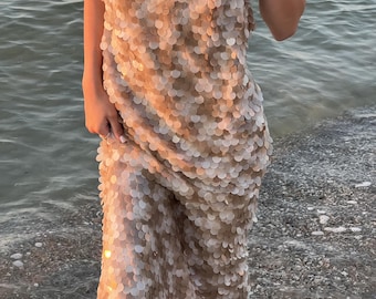 May include: A beige sequin dress with a halter neckline. The dress is knee-length and made of small, round sequins. The dress is worn near the water, with the waves gently lapping at the hem.