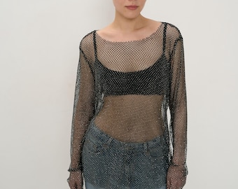 May include: A black mesh long-sleeve top with a scoop neck. The top is worn over a black bra and paired with light blue jeans. The mesh top has a sparkly, glittery appearance.