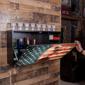 May include: A black wall-mounted bar cabinet with an American flag design on the drop-down door. The cabinet is stocked with liquor bottles and glassware. The bar is mounted on a wooden plank wall.