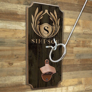 May include: A wooden wall-mounted game board with a dark brown finish, featuring a shield design with the letter "S" and the name "SIMPSON". It includes a bottle opener and a ring toss game. The ring is silver.