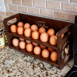 May include: A dark brown wooden egg holder with two tiers, filled with brown eggs. The holder has handles and the text "Teresa's Kitchen" and "Farm Fresh Eggs" is engraved on the wood. The holder sits on a speckled countertop.