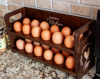 Pode incluir: Um suporte de madeira castanha escura para ovos com dois níveis, cheio de ovos castanhos. O suporte tem pegas e o texto "Teresa's Kitchen" e "Farm Fresh Eggs" está gravado na madeira. O suporte está sobre uma bancada salpicada.
