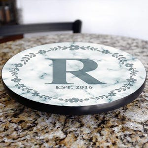 May include: Round marble-look decorative tray with a black edge. The tray features a large gray letter "R" in the center, surrounded by a floral wreath. The text "EST. 2016" is below the letter.