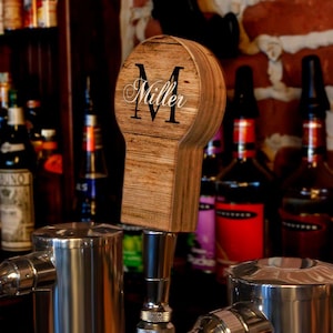 May include: A wooden tap handle with the name "Miller" and a large "M" in black script. The handle is made of light brown wood and is set against a blurred background of liquor bottles and a brick wall. The tap handle is attached to a stainless steel tap.