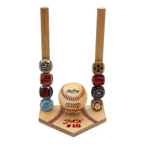 May include: A baseball-themed display stand with a baseball and several championship rings. The stand has two wooden uprights and a wooden base shaped like home plate. The baseball is cream-colored with red stitching. The rings are gold, red, and blue.