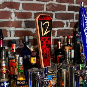 May include: A wooden tap handle with a red and black coffin-shaped design. The number 12 is prominently displayed in white. The handle features a vine and leaf pattern. Various liquor bottles are visible in the background, with a brick wall.