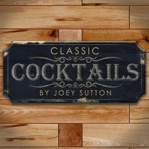 May include: A rectangular wooden sign with the words "CLASSIC COCKTAILS BY JOEY SUTTON" in a vintage font. The sign is dark blue with a distressed finish, mounted on a wooden plank background.