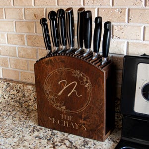 May include: A wooden knife block with a floral wreath design and the letter "n" engraved in the center. The block holds several knives with black handles. The text "THE McCRAY'S" is engraved below the wreath.