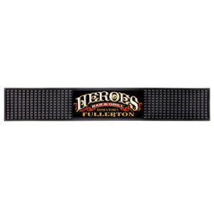 May include: A black bar mat with a textured surface and the words "HEROES BAR & GRILL DOWNTOWN FULLERTON" in gold and red lettering. The mat is rectangular and designed to catch spills.