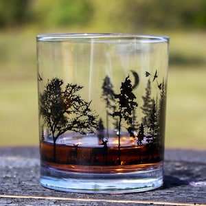 May include: A clear rocks glass with a black forest scene etched on the side. The design includes trees, deer, birds, and a crescent moon. The glass is filled with a brown liquid, likely a spirit, and sits on a wooden surface.