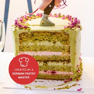 Pode incluir: Um bolo de vários andares verde e rosa com cobertura branca e pétalas de rosa no topo. Uma faca está cortando o bolo. O bolo está etiquetado como "Criado por um mestre pasteleiro alemão".