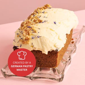 Könnte beinhalten: Ein Brotkuchen, der mit weißem Frosting überzogen und mit Walnüssen und blauen essbaren Blüten verziert ist. Der Kuchen steht auf einem klaren Glaskuchenständer. Der Text "CREATED BY A GERMAN PASTRY MASTER" befindet sich auf einem roten Kreis mit einem Kochmützen-Symbol.