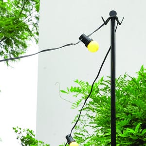May include: A black metal pole with a string of yellow lights hanging from it. The lights are strung across a yard with green trees in the background.