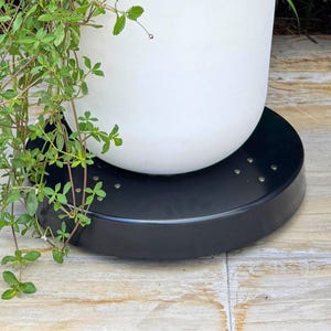 May include: A white ceramic planter sits on a black, circular plant stand. The stand has small, evenly spaced holes. Green leafy vines cascade down from the top of the planter, creating a natural, decorative display.