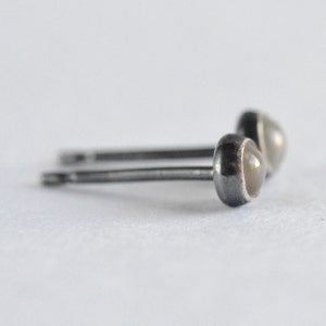 May include: A pair of small stud earrings. Each earring features a round, light-colored stone set in a dark metal bezel. The posts are thin and silver-toned. The earrings are displayed on a white background.