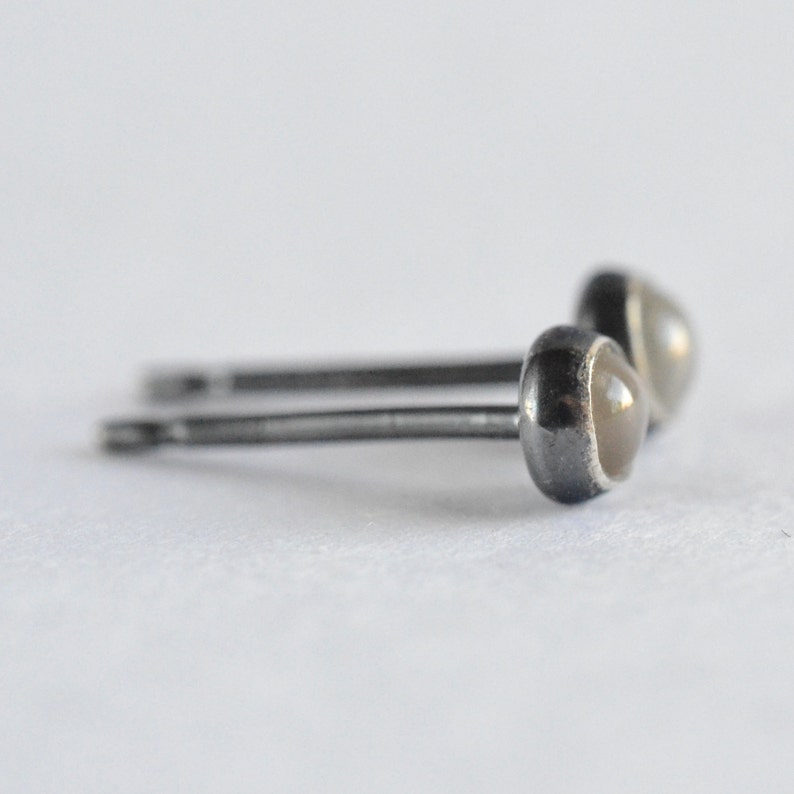 May include: A pair of small stud earrings. Each earring features a round, light-colored stone set in a dark metal bezel. The posts are thin and silver-toned. The earrings are displayed on a white background.