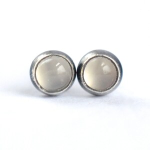 May include: A pair of stud earrings with a silver-tone metal setting. Each earring features a round, pale gray stone. The earrings are displayed against a white background, highlighting their simple design.