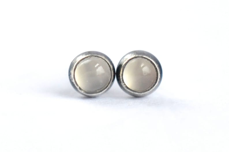 May include: A pair of stud earrings with a silver-tone metal setting. Each earring features a round, pale gray stone. The earrings are displayed against a white background, highlighting their simple design.