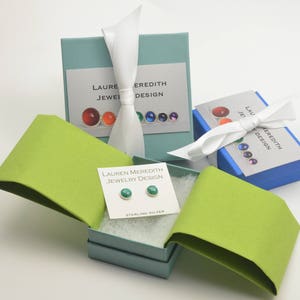 May include: An image of jewelry packaging featuring small, teal stud earrings on a white card labeled "STERLING SILVER." The earrings are displayed in an open, light blue box with a lime green lid. Other boxes with white ribbons and the text "LAUREN MEREDITH JEWELRY DESIGN" are also visible.