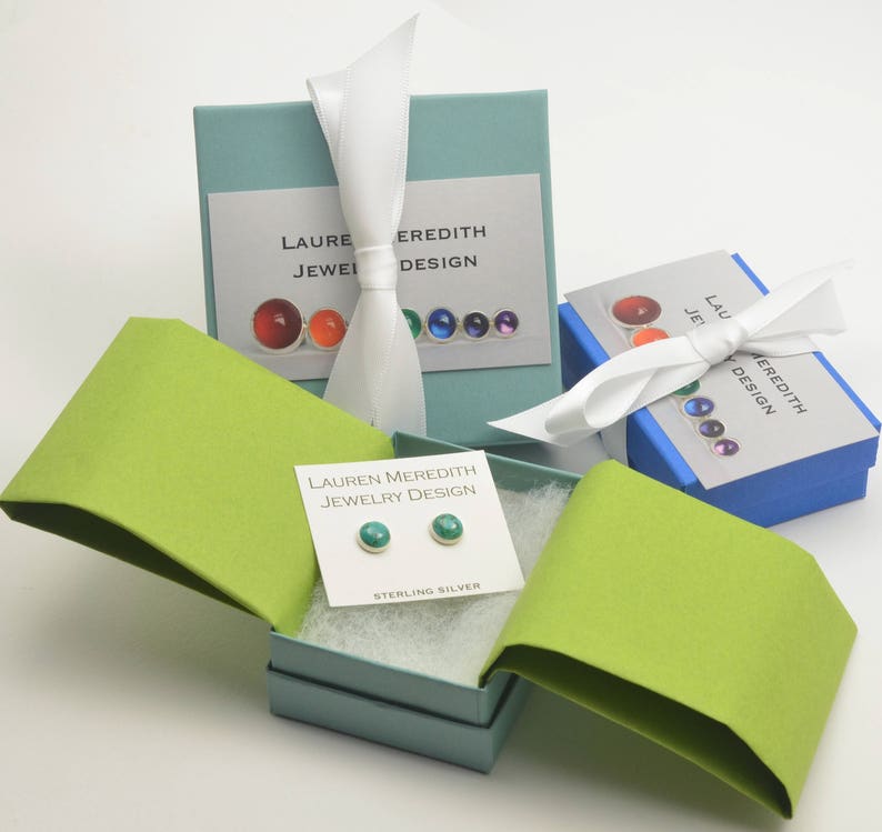 May include: An image of jewelry packaging featuring small, teal stud earrings on a white card labeled "STERLING SILVER." The earrings are displayed in an open, light blue box with a lime green lid. Other boxes with white ribbons and the text "LAUREN MEREDITH JEWELRY DESIGN" are also visible.