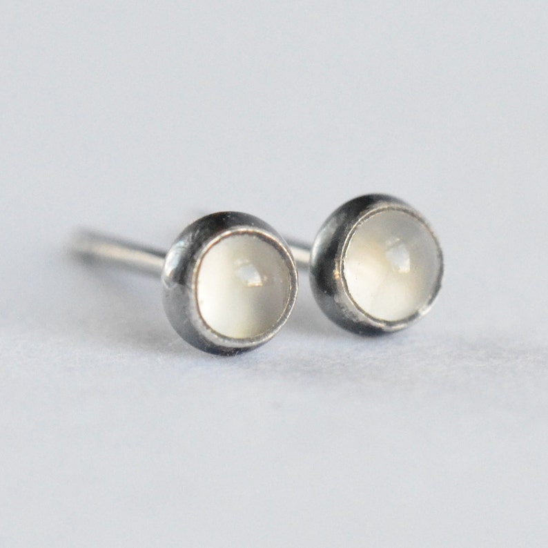 May include: A pair of stud earrings featuring a round, off-white stone set in a dark metal bezel. The earrings have a simple, minimalist design with a post backing. The stones have a subtle, pearlescent sheen, and the metal has a slightly oxidized finish.