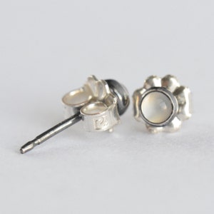 May include: A pair of silver stud earrings with a flower-shaped setting. Each earring features a small, round, off-white stone. The post and backings are visible, showing the details of the jewelry's construction.