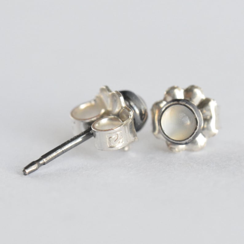May include: A pair of silver stud earrings with a flower-shaped setting. Each earring features a small, round, off-white stone. The post and backings are visible, showing the details of the jewelry's construction.