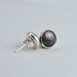 May include: A pair of silver stud earrings with round, dark gray, and silver-flecked stones. The earrings have a polished finish and are set in a simple bezel setting. The earring posts are visible, with a 925 stamp.