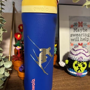 May include: A blue reusable water bottle with a yellow lid and a gold sticker of a skier. The sticker says "owala" in pink letters. The bottle is on a wooden surface.