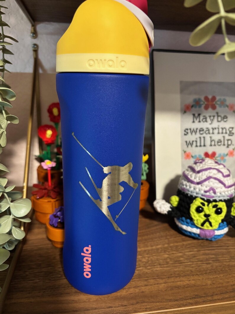 May include: A blue reusable water bottle with a yellow lid and a gold sticker of a skier. The sticker says "owala" in pink letters. The bottle is on a wooden surface.