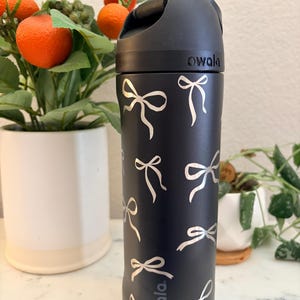 Puede incluir: Botella de agua aislada negra con tapa abatible y estampado de lazos plateados. La botella tiene el nombre de la marca "owala" impreso. La botella es cilíndrica y parece estar hecha de acero inoxidable.