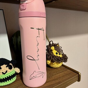 May include: Pink water bottle with a black dolphin design and the name "Finny" written in cursive on the side.