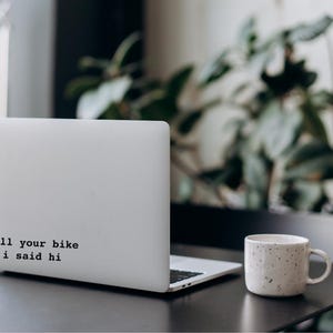 Peut inclure: Un ordinateur portable blanc avec un autocollant noir qui dit "tell your bike i said hi" sur le couvercle. L'ordinateur portable est pos&eacute; sur une table noire avec une tasse &agrave; caf&eacute; blanche.