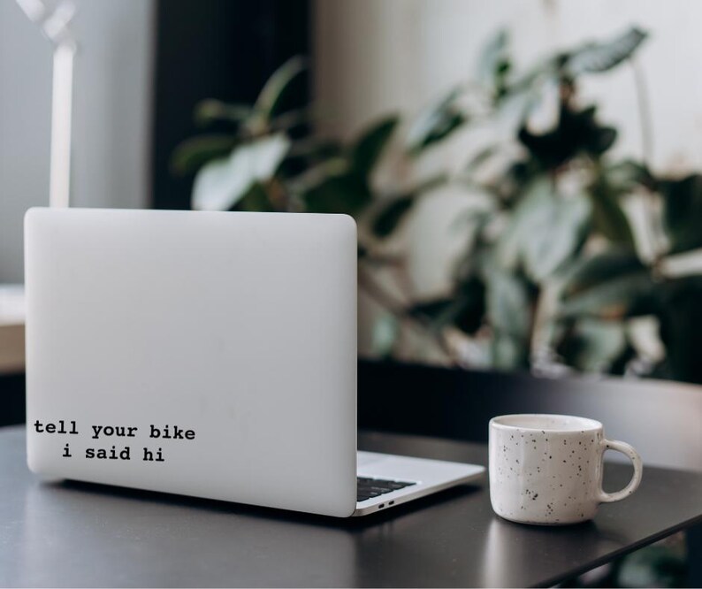 Peut inclure: Un ordinateur portable blanc avec un autocollant noir qui dit "tell your bike i said hi" sur le couvercle. L'ordinateur portable est pos&eacute; sur une table noire avec une tasse &agrave; caf&eacute; blanche.