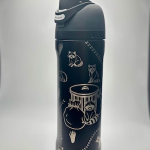 Puede incluir: Botella de agua aislada negra con un diseño de mapache y contenedor de basura. La botella tiene una tapa negra con una etiqueta blanca "to unlock".