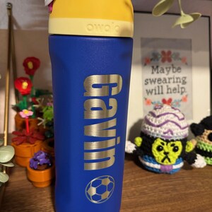 May include: A blue water bottle with a yellow lid and a silver nameplate that says "Gavin". A soccer ball is printed on the bottle. The bottle is sitting on a wooden shelf.