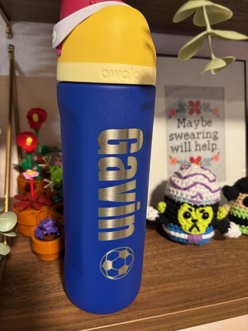 May include: A blue water bottle with a yellow lid and a silver nameplate that says "Gavin". A soccer ball is printed on the bottle. The bottle is sitting on a wooden shelf.