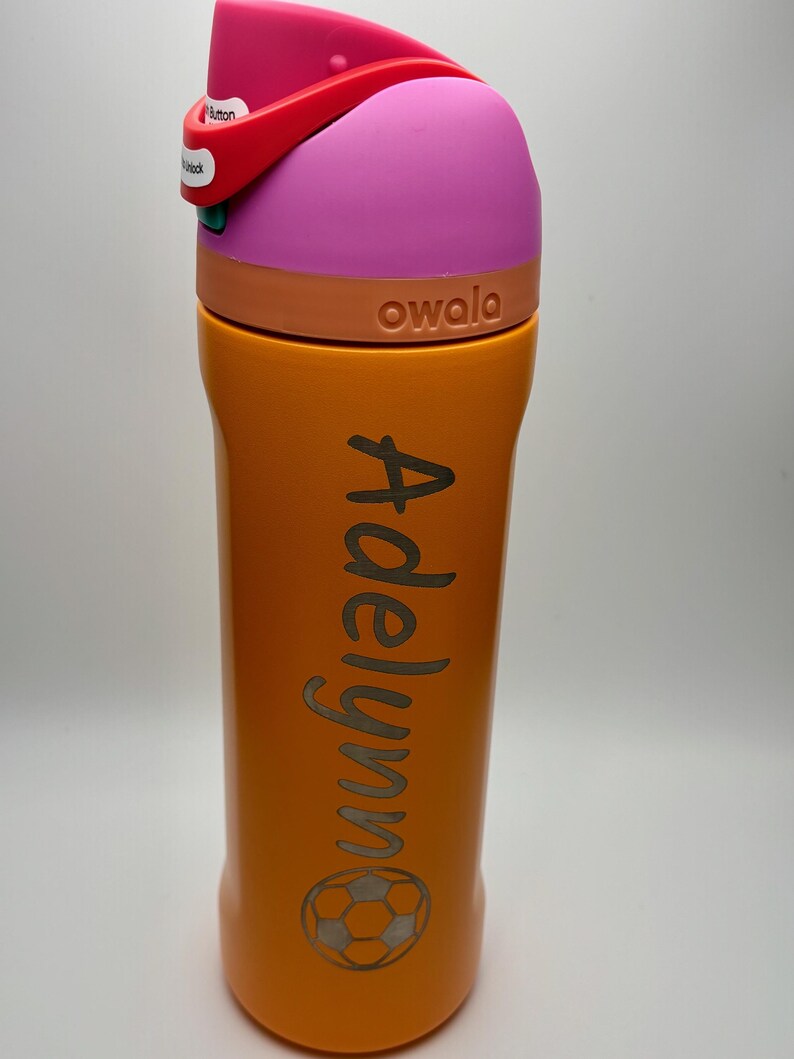 May include: Orange and pink reusable water bottle with a soccer ball design and the name "Adelynn" engraved on the side. The bottle is made by the brand "owala".