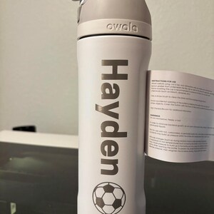 May include: White water bottle with a gray lid and a soccer ball design. The bottle has the name "Hayden" printed on it and the word "owala" on the lid.