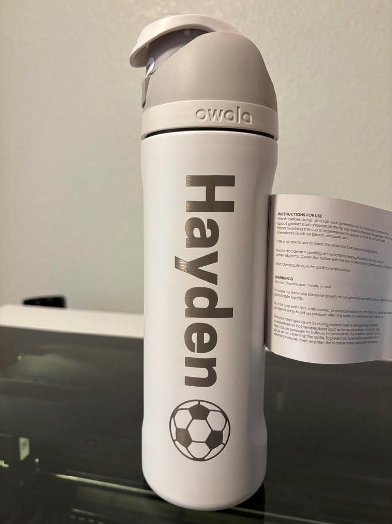 May include: White water bottle with a gray lid and a soccer ball design. The bottle has the name "Hayden" printed on it and the word "owala" on the lid.